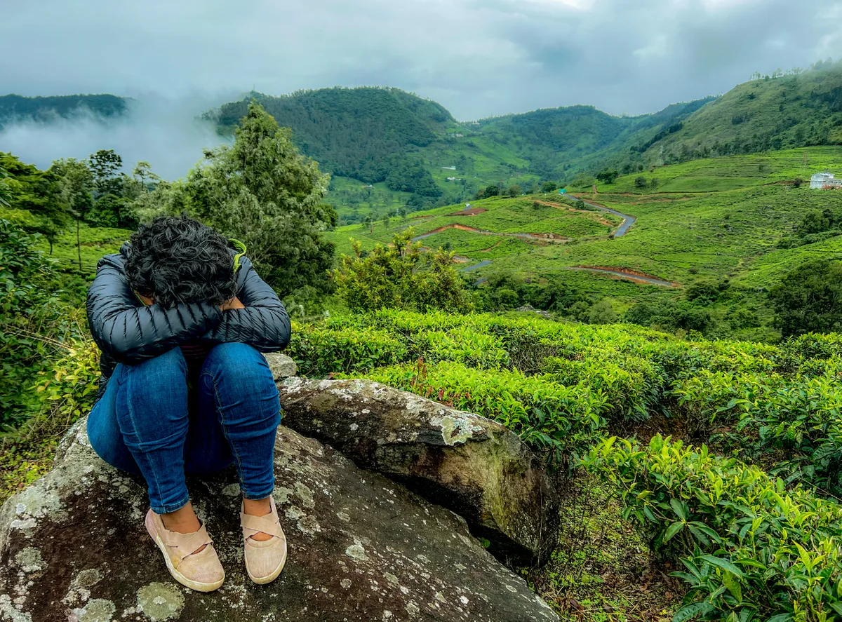 A figure sitting on a rock with her face hidden in her arms, tea hills beyond.