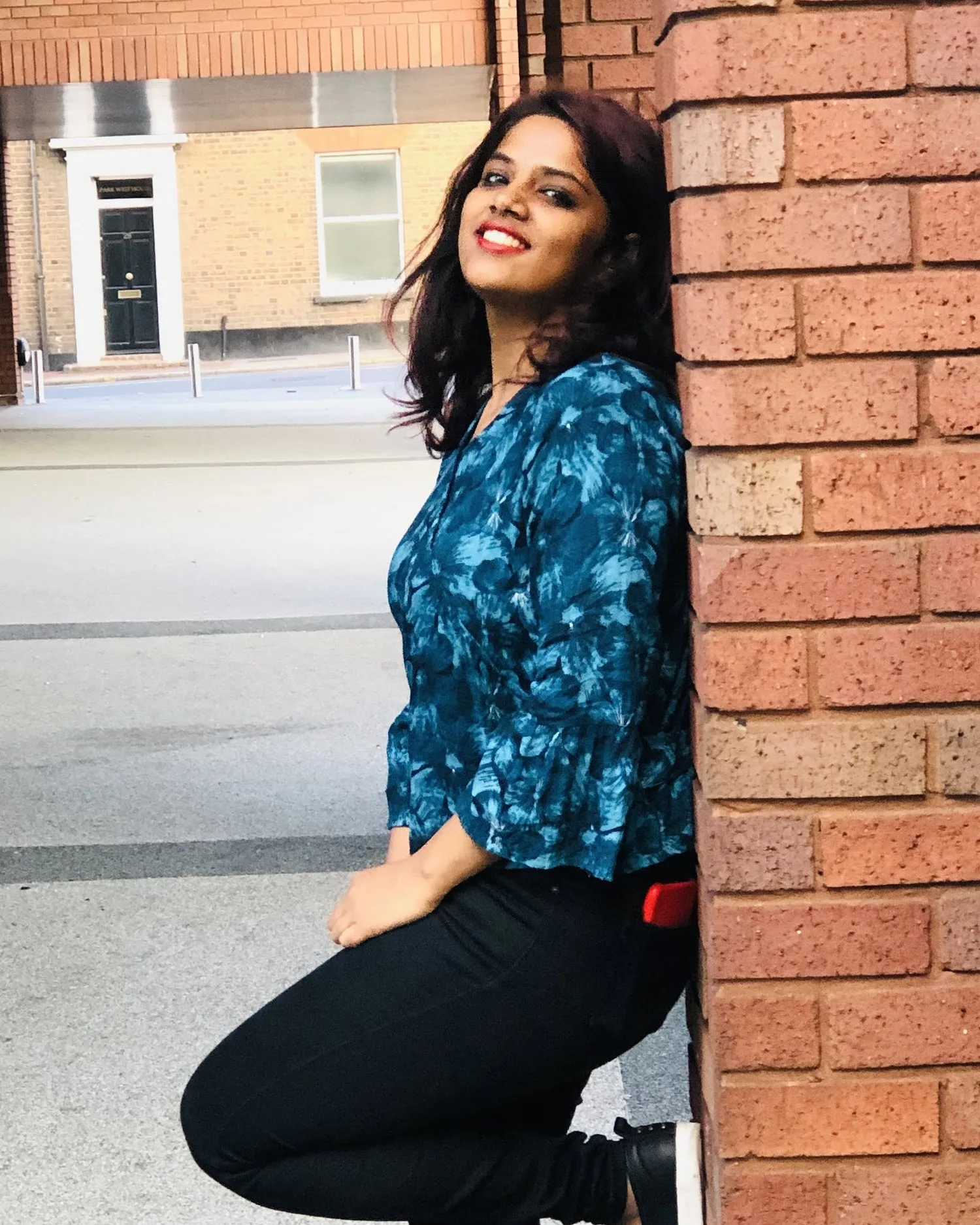 A woman leaning against a brick pillar in soft daylight, smiling.