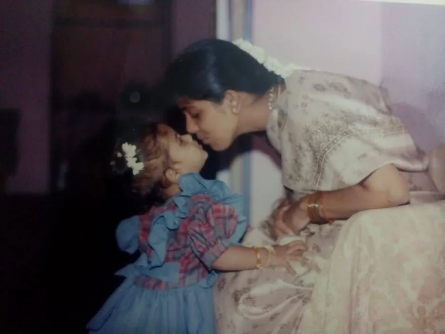 An old photograph: a mother in a pale saree leaning down to kiss her small daughter.