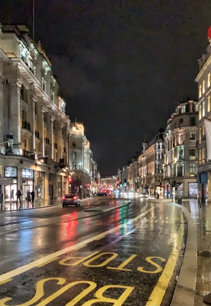 A rain-slicked London street at night, a bus stop reflected in the wet road.
