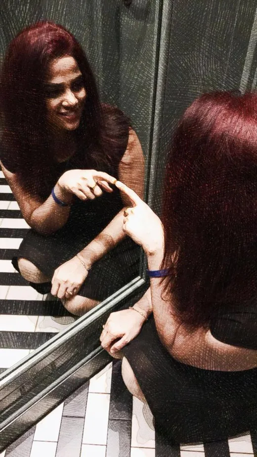 A woman crouched laughing in front of a large mirror, her reflection beside her.