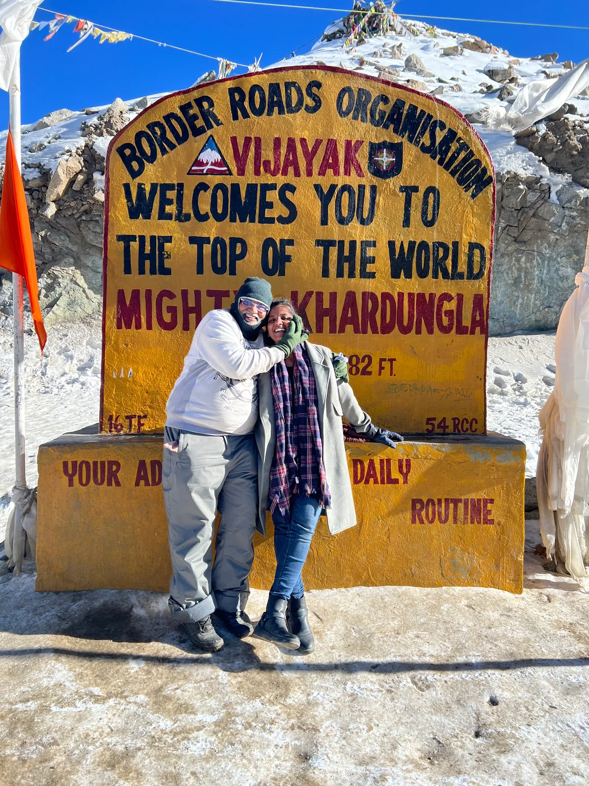 A father and daughter holding each other in front of a painted signboard at a snowy mountain pass.