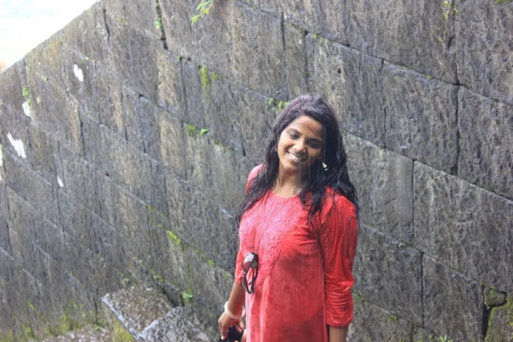 A young woman in a red kurta, wet from the rain, smiling against a stone wall.