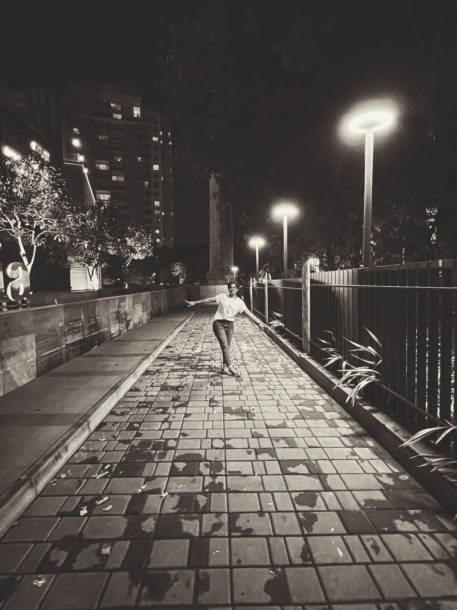 A lone figure walking a lamplit boardwalk in black and white.