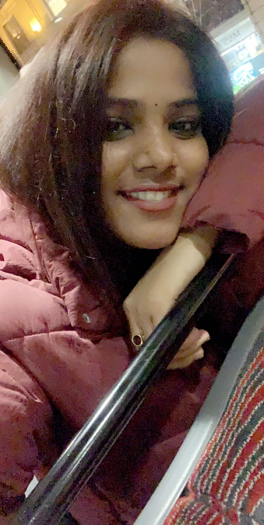 A young woman smiling on a night bus, resting her chin on the seat rail.