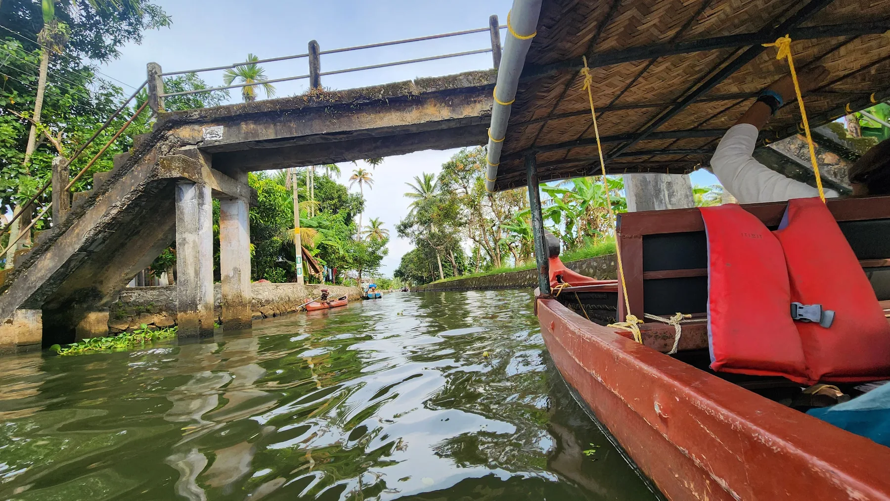 A small red boat on green backwaters, beneath a bridge.